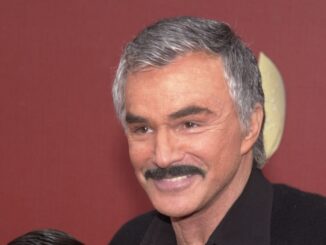 Burt Reynolds smiling in front of a brown backround