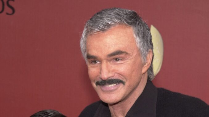 Burt Reynolds smiling in front of a brown backround