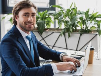 A handsome man sat at a desk wokring on a laptop smiling. real God of war