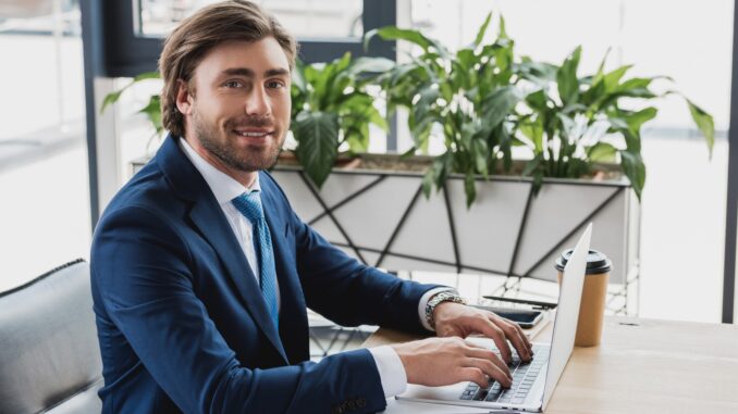 A handsome man sat at a desk wokring on a laptop smiling. real God of war