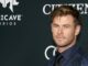 Chris Hemsworth in a black suit and shirt, showing no sign of having been inflated