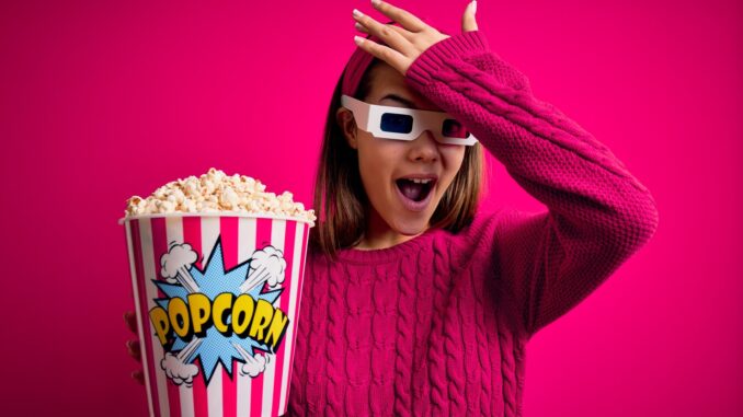 beautiful girl watching movie using 3d glasses eating box with popcorns surprised with hand on head after seeing movie blooper