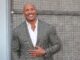 Dwayne Johnson in a grey jacket leaning against a wall. Vin Diesel and The Rock
