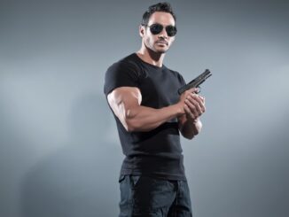 Action hero muscled man holding a gun. Wearing black t-shirt with pants and sunglasses. Studio shot against grey. MAverick cop