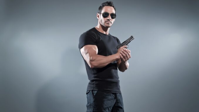 Action hero muscled man holding a gun. Wearing black t-shirt with pants and sunglasses. Studio shot against grey. MAverick cop