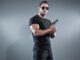 Action hero muscled man holding a gun. Wearing black t-shirt with pants and sunglasses. Studio shot against grey. MAverick cop