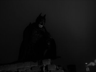 A man dressed as a bat in mask, helmet, armor and black cloak sits at the top of the building against the background of night lights and sky. Cosplay. The Batman 2