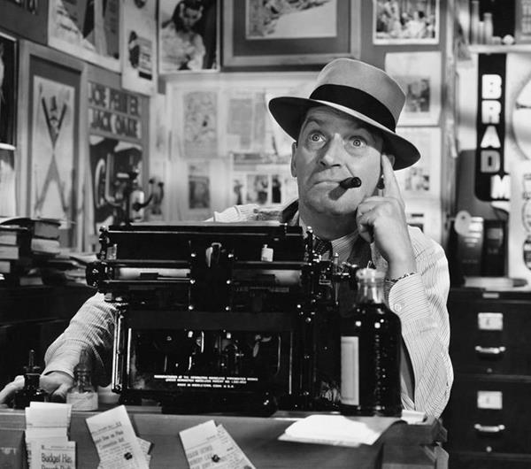 1950s man in a hat sitting in front of a typewriter with a quizzical look